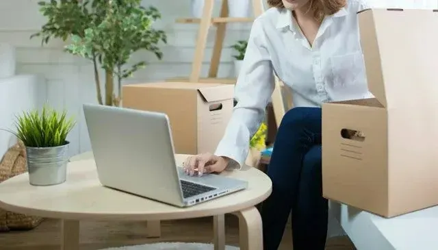 Een laptop op een houten tafel, naast een plant in een zilveren pot. Kartonnen dozen en een houten ladder op de achtergrond.