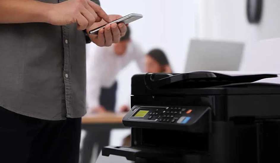 Een kantooromgeving met een zwarte printer op de voorgrond. Iemand gebruikt een smartphone, op de achtergrond zijn mensen aan het werk.
