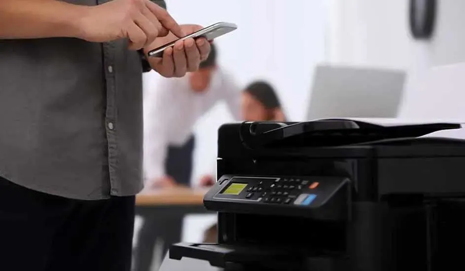 Een kantooromgeving met een zwarte printer op de voorgrond. Iemand gebruikt een smartphone, op de achtergrond zijn mensen aan het werk.