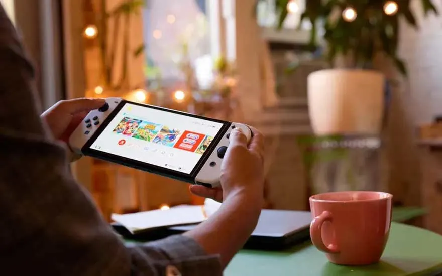 Une console de jeu blanche à écran est tenue à deux mains. Une tasse rose est sur une table verte, près d'une plante.