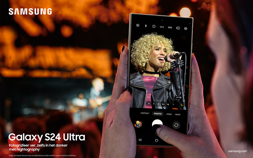 Handen houden een Samsung telefoon vast. Op het scherm staat een zangeres met een microfoon op een podium met oranje licht.