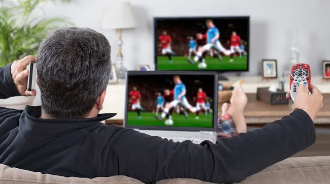 Een tv en laptop tonen een voetbalwedstrijd. Er is een rode afstandsbediening in de hand.