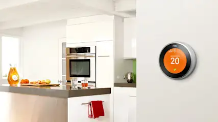 Dans une cuisine blanche, un thermostat Nest rond affiche 20 degrés Celsius sur un mur blanc. Sur le comptoir, il y a des oranges et un pichet de jus.