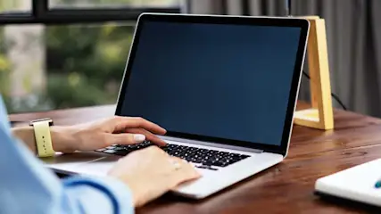 Laptop with a dark screen on a wooden desk. A notepad and pen are to the side. Window in background.