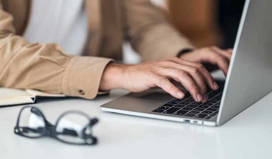 Une personne tape sur un ordinateur portable gris sur une table blanche. Des lunettes et un cahier sont à gauche.