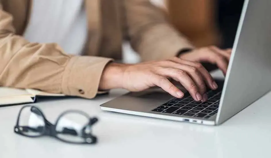 Iemand typt op een laptop op een witte tafel. Een notitieboek en een bril liggen ernaast.