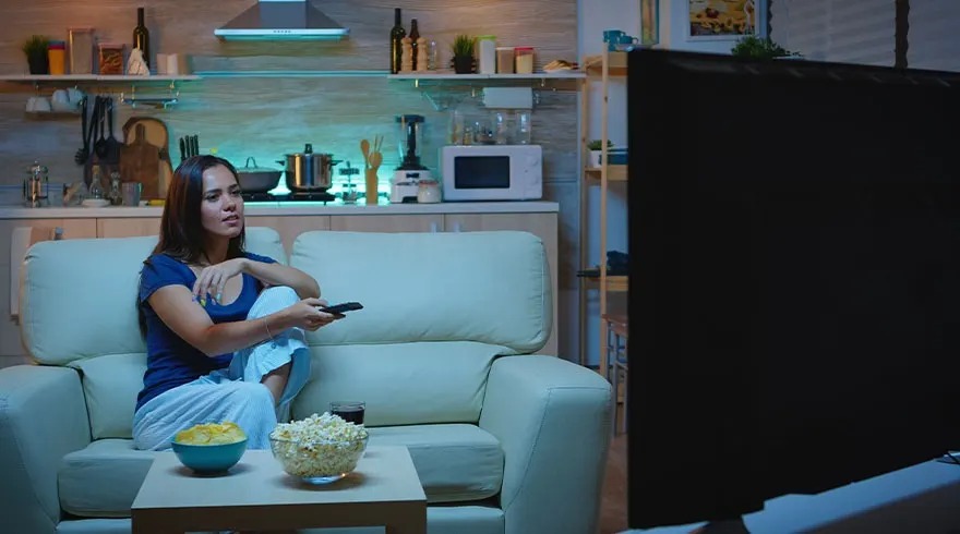 Femme assise sur canapé beige dans cuisine ouverte regarde la TV, télécommande, popcorn et chips prêts pour la soirée film.
