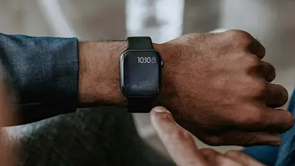 Een zwart horloge toont 10:10 op een pols met een vinger die het scherm aanraakt, tegen een vage achtergrond.