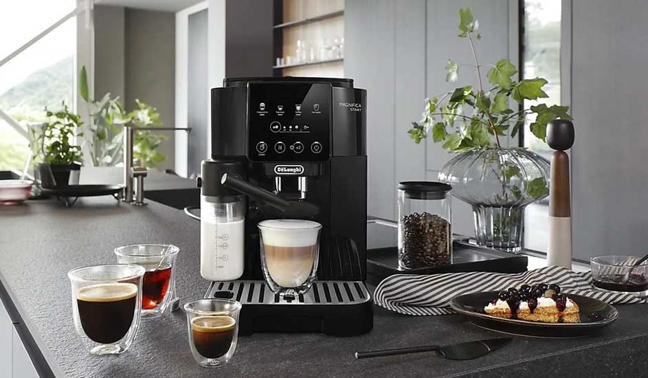 A black automatic coffee machine stands on a kitchen worktop, surrounded by cups, glasses, milk, coffee beans and pastries in a modern kitchen.