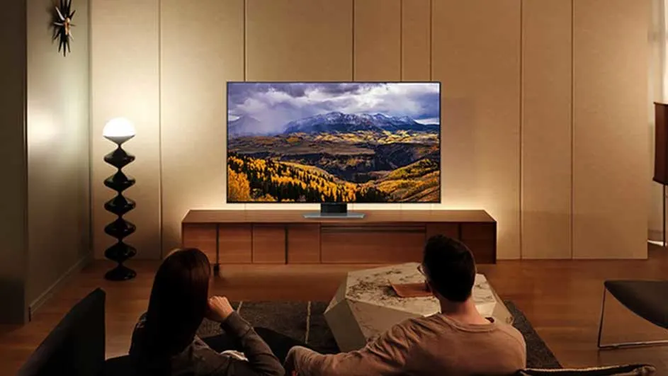 Un couple est assis dans un salon chaleureux et regarde une télé montrant un paysage de montagne sur un meuble épuré.