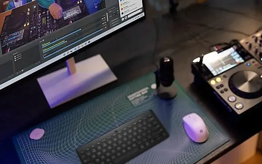 Une table avec un écran, un clavier noir, une souris blanche, un microphone et une table de mixage DJ.