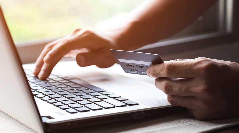 A laptop sits on a surface, with a credit card held near the keyboard. Fingers type on the keys, suggesting a transaction or online process.
