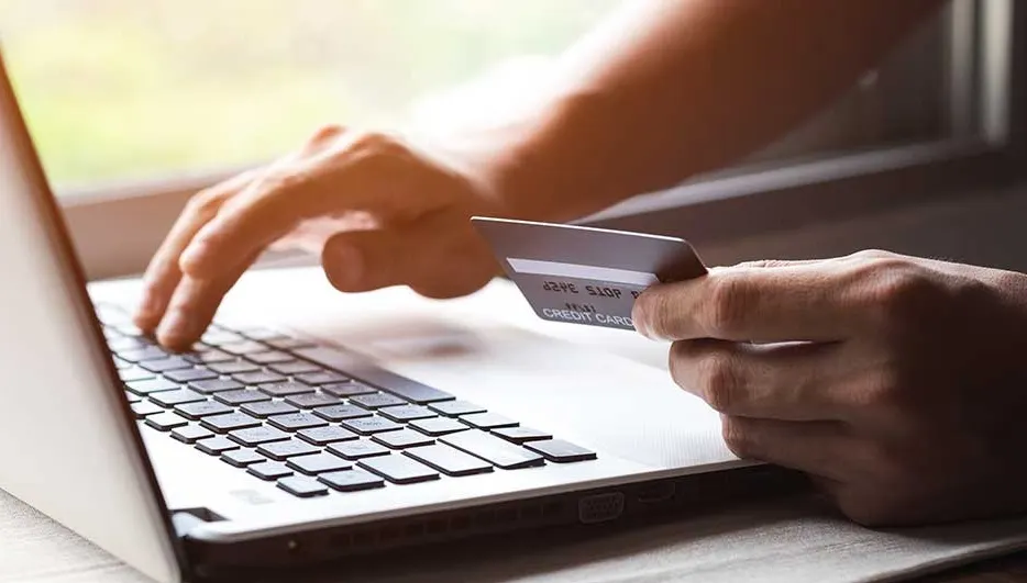 A laptop sits on a surface, with a credit card held near the keyboard. Fingers type on the keys, suggesting a transaction or online process.