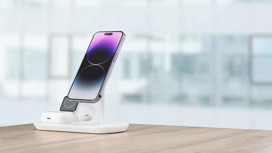 Un téléphone violet sur un support de charge blanc, avec des écouteurs et une montre, sur une table en bois.