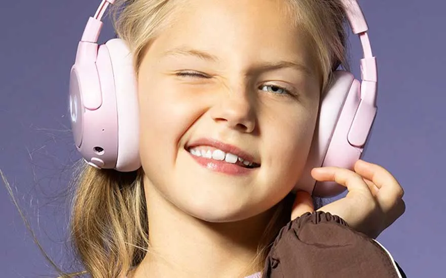 Een jong meisje met roze koptelefoon knipoogt speels naar de camera, genietend van muziek terwijl ze breed glimlacht tegen een paarse achtergrond.