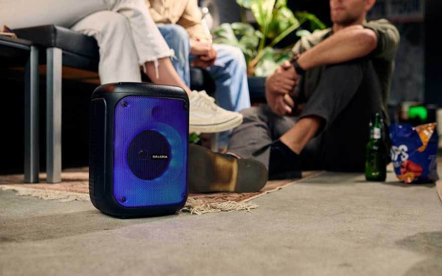 A blue Salora speaker sits on a rug. In the background are people, a bottle, and a bag of chips.