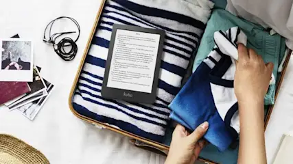 Een koffer gevuld met kleding, een Kobo e-reader, foto's en een paspoort op een witte achtergrond.