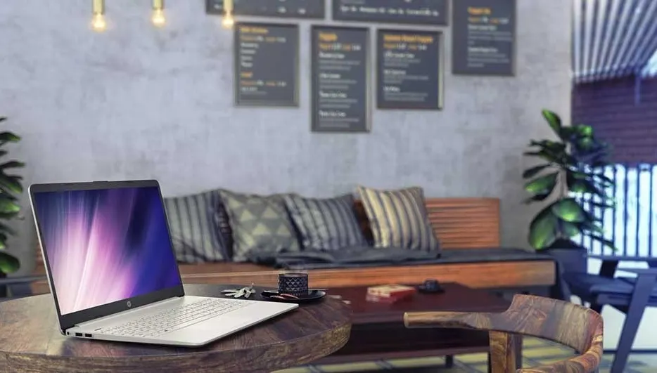 Laptop on a wooden table. Behind, a couch with pillows and wall menus. Plant and blinds visible.