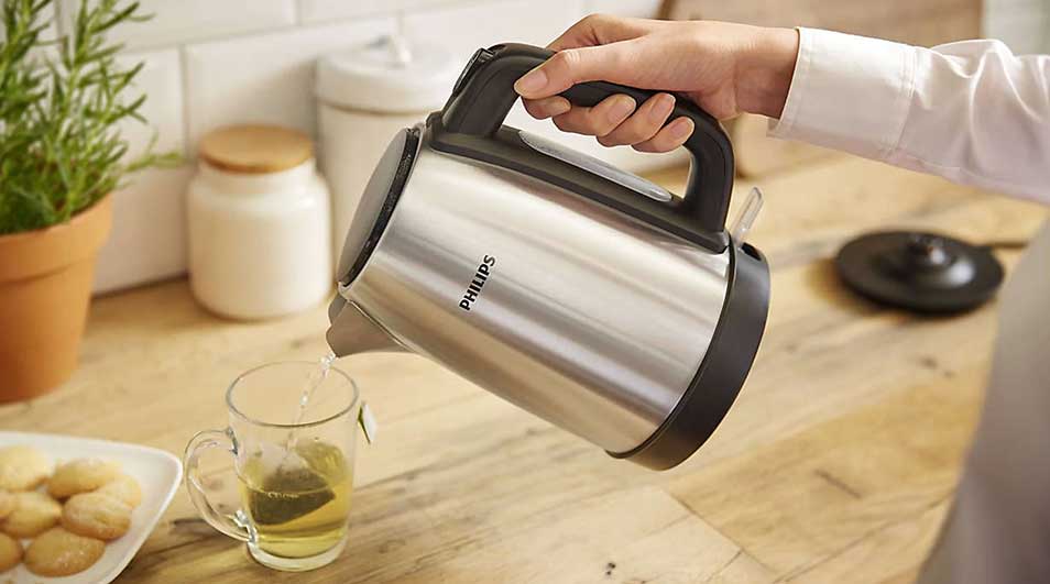 Une bouilloire Philips en métal verse de l'eau chaude dans une tasse à thé sur une table en bois, près d'une plante et de biscuits.