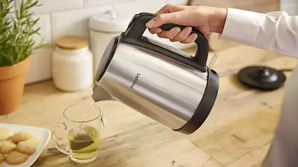 Zilveren waterkoker schenkt water in een glas met theezakje op een houten aanrecht. Koekjes en een plant zijn ook zichtbaar.