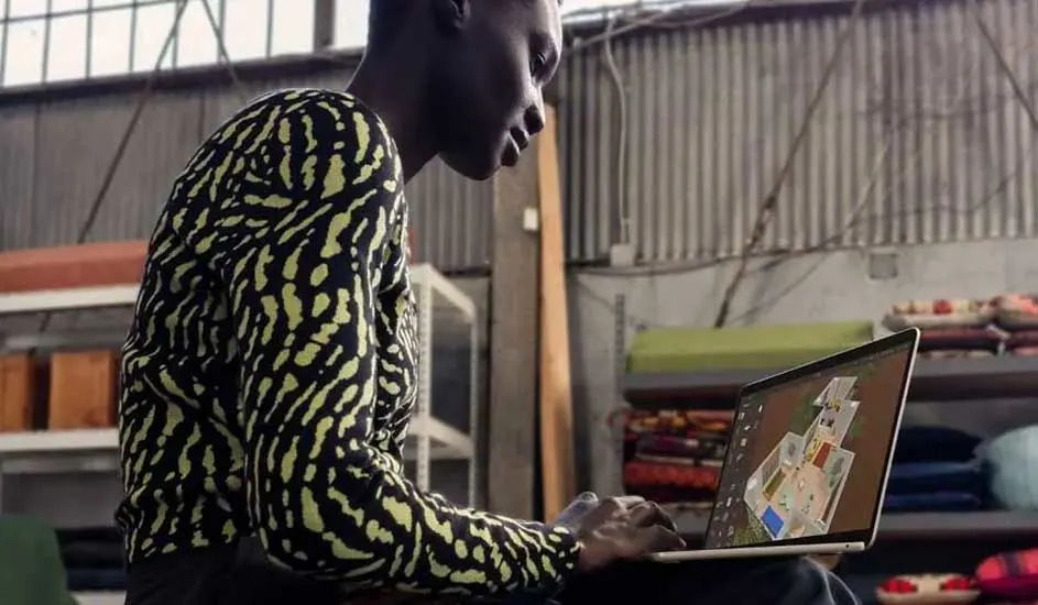 Iemand zit in een industriële ruimte met een laptop waarop een 3D-plattegrond staat, omgeven door rekken en gestapelde stoffen.