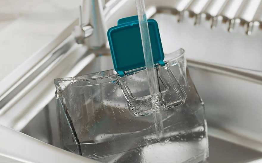 De l'eau coule d'un robinet dans un récipient transparent avec un couvercle bleu-vert dans un évier en acier inoxydable.