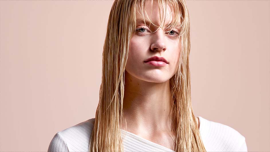 Portrait d'une personne aux cheveux longs et raides, portant un haut blanc sur fond uni.