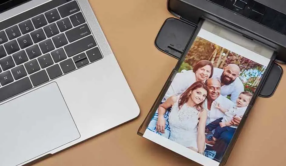 Un ordinateur portable argenté est à côté d'une imprimante noire. Une photo de famille est en cours d'impression. Fond beige.