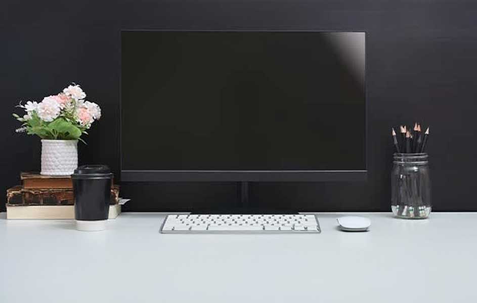Bureau met een zwarte monitor, wit toetsenbord en muis. Een vaas met bloemen en potloodhouder staan ernaast.