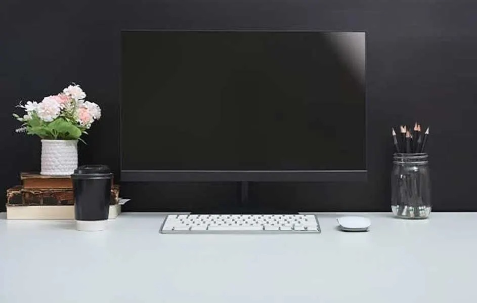 Bureau met een zwarte monitor, wit toetsenbord en muis. Een vaas met bloemen en potloodhouder staan ernaast.