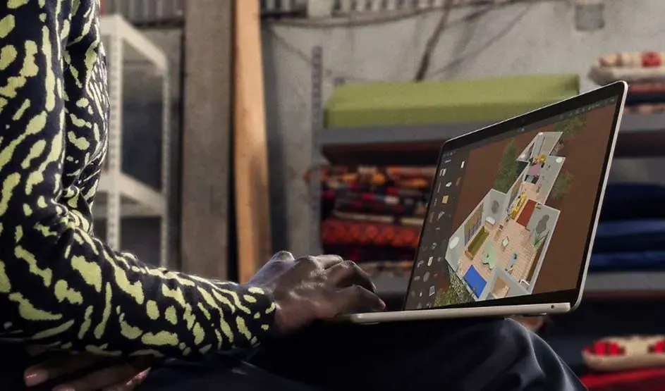 Iemand gebruikt een dunne laptop op schoot in een creatieve ruimte; op het scherm staat een kleurrijk ontwerp van een plattegrond.