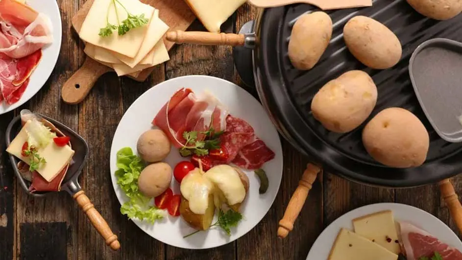 Vue de dessus d'une table en bois avec raclette, pommes de terre, fromage, charcuterie et légumes.