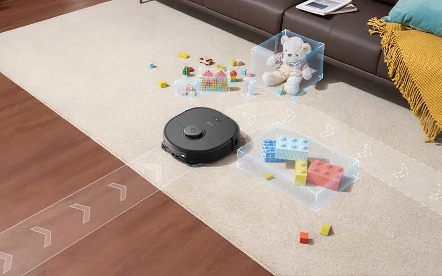 Een zwarte robotstofzuiger op een beige tapijt met speelgoed, waaronder een teddybeer en bouwstenen, tegen een achtergrond van houten vloer en een bruine sofa.