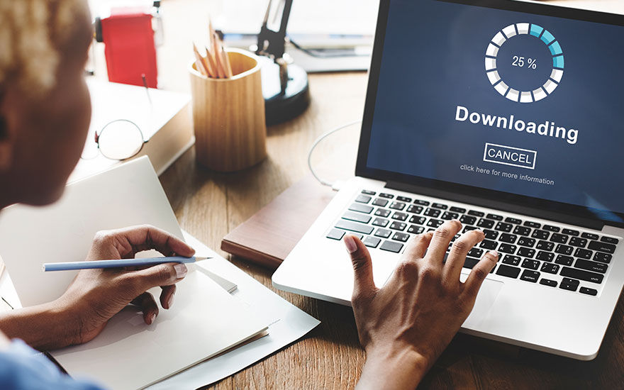 Iemand typt op een laptop met een downloadscherm (25%) en houdt een potlood boven een notitieboek op een houten bureau.