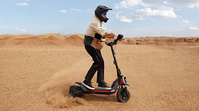 Een zwarte scooter met rode details rijdt door een zandlandschap onder een blauwe lucht met wolken.