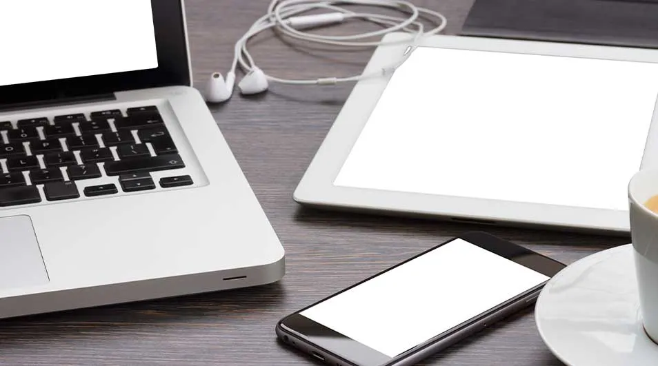 Sur une table en bois, un ordinateur portable, une tablette, un smartphone, des écouteurs et une tasse blanche.