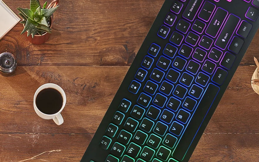 Vue du dessus d'un bureau en bois avec un clavier rétroéclairé, une tasse de café, une plante et un petit récipient métallique.