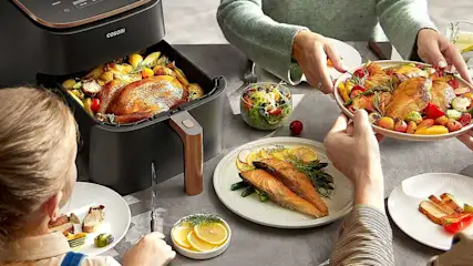 Een luchtfriteuse naast borden met kip, vis en groenten op een grijze tafel. Een kom met salade staat ernaast.