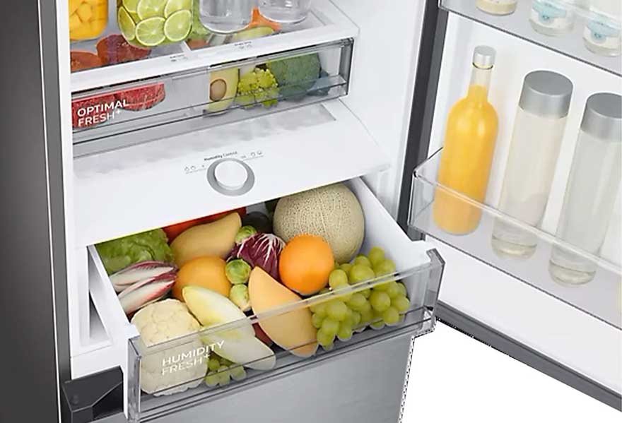 Een open koelkast gevuld met fruit, groenten en flessen. De lades bevatten meloenen, sinaasappels en druiven.