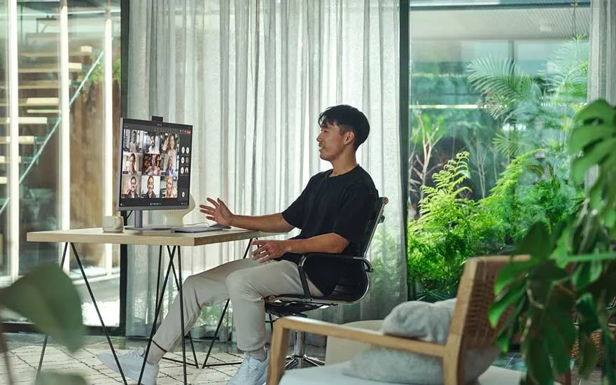 Een man voert vanuit een groene, huiselijke ruimte een videogesprek aan een kleine werktafel met monitor en comfortabele bureaustoel.