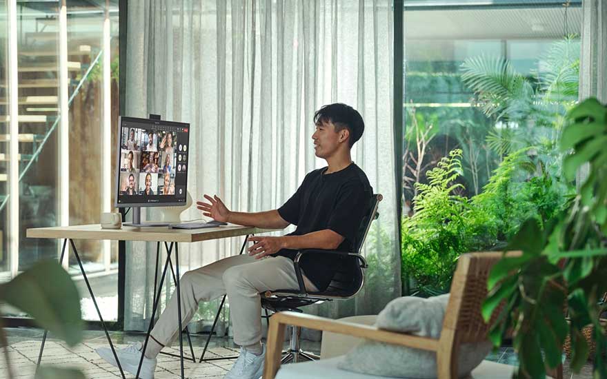 Un homme participe à une visioconférence dans une pièce verdoyante, installé à un petit bureau avec écran et fauteuil confortable.