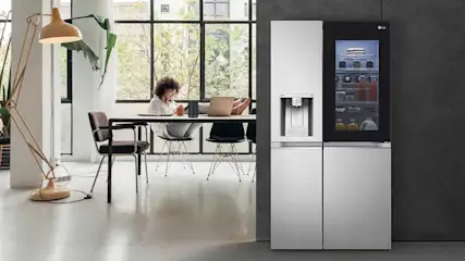 Een persoon werkt aan tafel bij grote ramen, terwijl rechts een moderne koelkast met waterdispenser en verlicht scherm staat.
