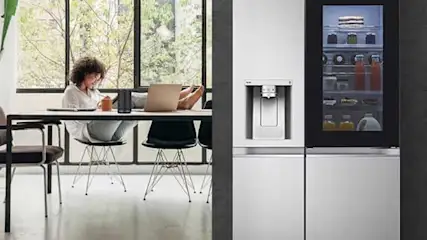 A person works at a table by large windows, while a modern fridge with a water dispenser and lit screen stands on the right.