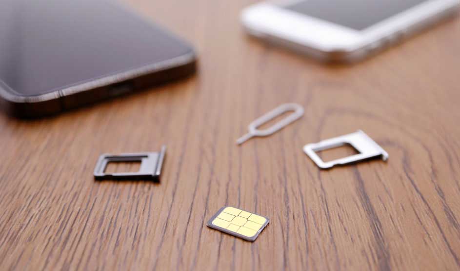 Sur une table en bois, on voit deux smartphones, une carte SIM, deux supports de carte SIM et un outil d'extraction.