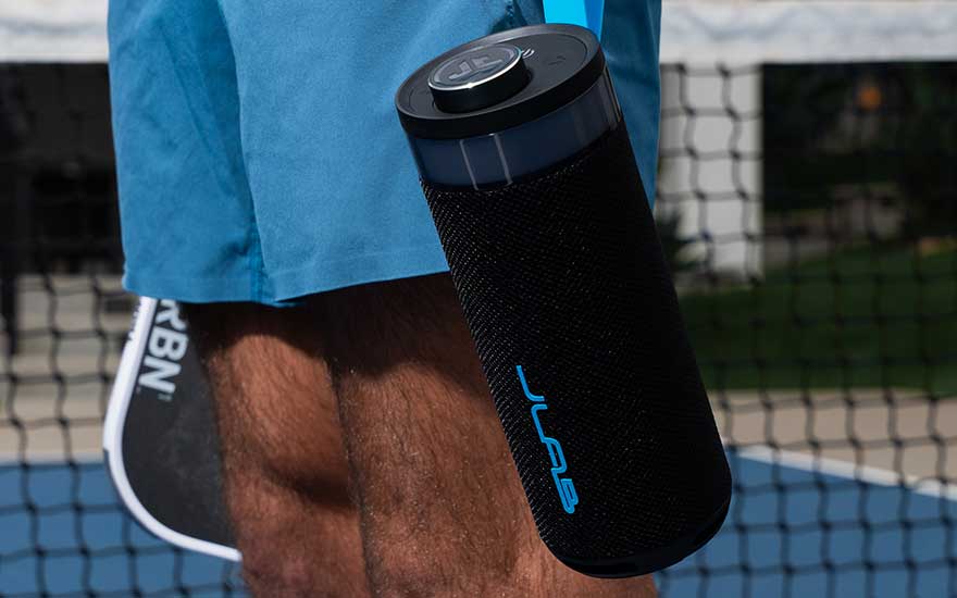 Zwarte draagbare bluetooth speaker met blauw logo, gedragen door iemand in blauwe sportkleding op een tennisbaan.