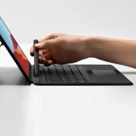 Een zwarte tablet met toetsenbord en standaard staat op een witte ondergrond. Een hand houdt een zwarte stylus vast.