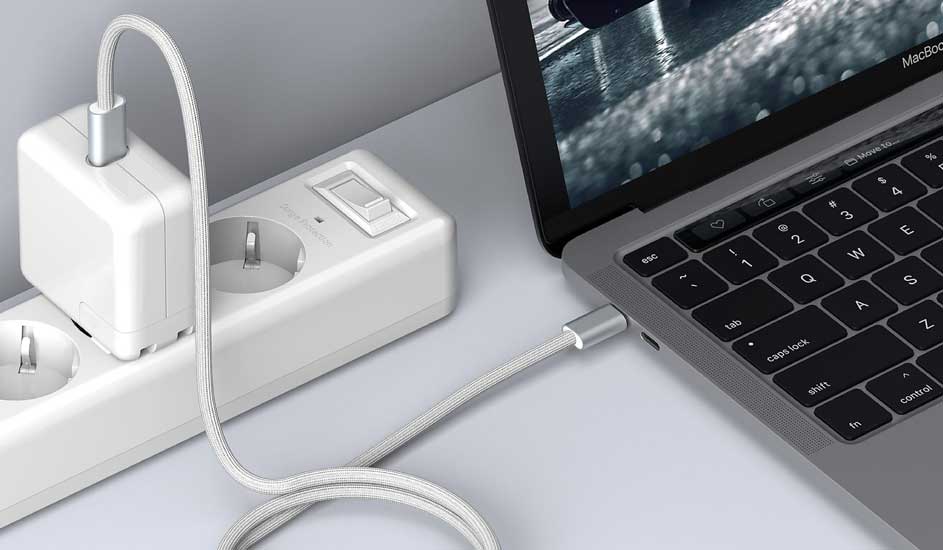 Un ordinateur portable gris est branché sur une prise murale blanche avec un câble tressé gris.