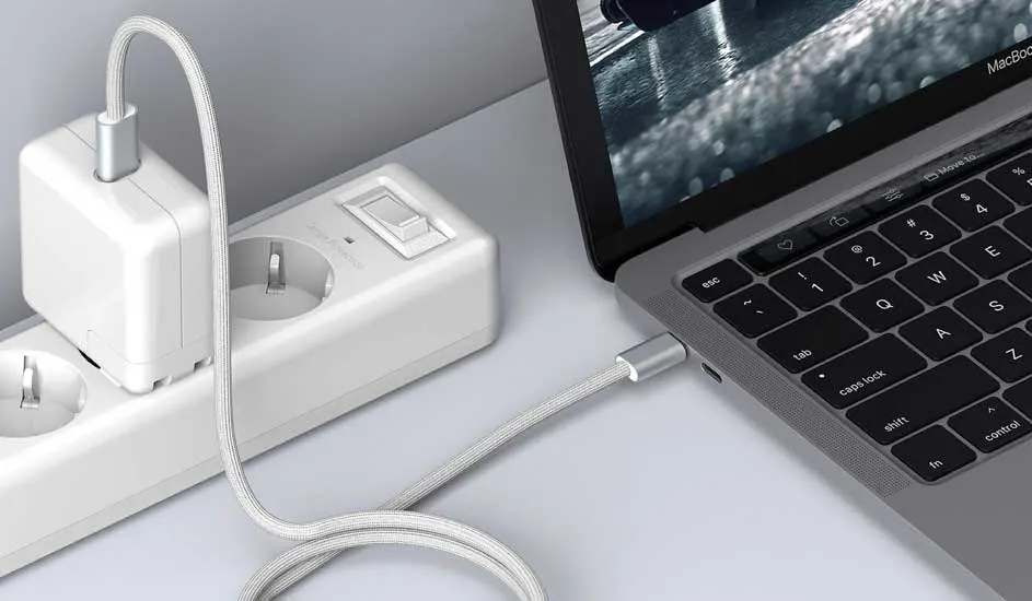 Un ordinateur portable gris est branché sur une prise murale blanche avec un câble tressé gris.