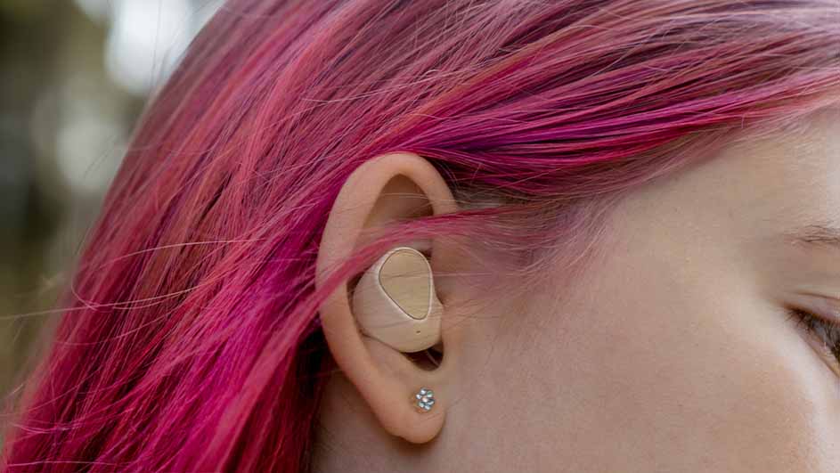 Oreillette beige dans une oreille, cheveux rose vif. Clou d'oreille brillant visible. Arrière-plan flou, nature.