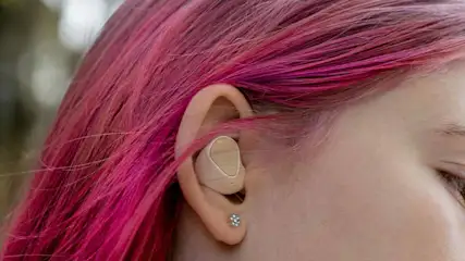 Oreillette beige dans une oreille, cheveux rose vif. Clou d'oreille brillant visible. Arrière-plan flou, nature.